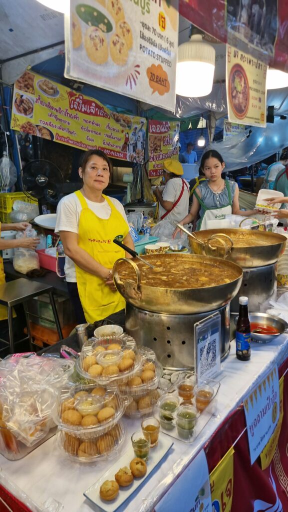 Vendor in a yellow apron in front of her woks of jay dishes
